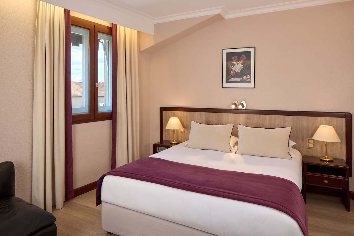 Une chambre d’hôtel avec un grand lit, deux tables de chevet avec des lampes, une fenêtre avec des rideaux et un tableau encadré au mur.