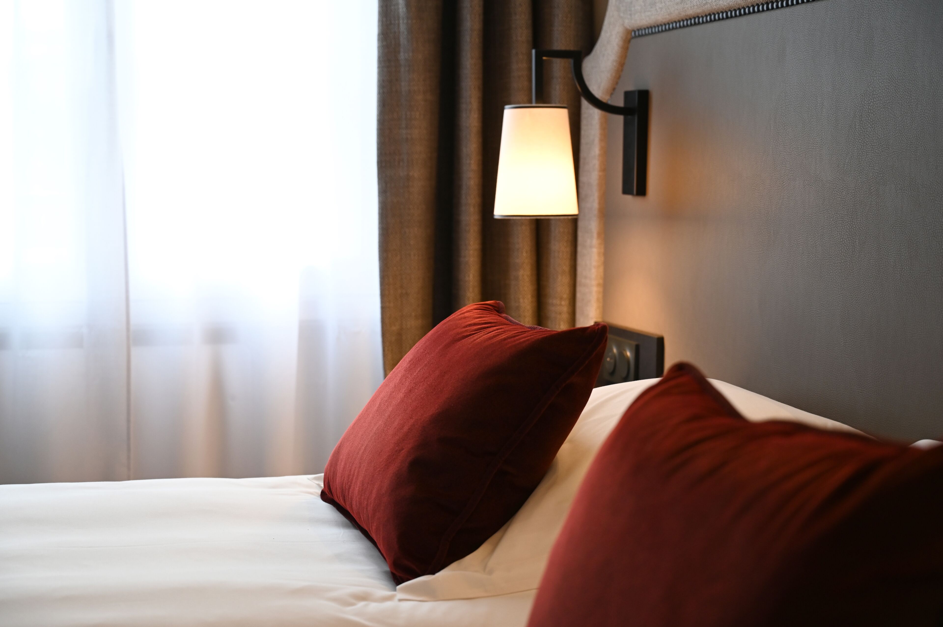Una habitación de hotel con una cama, almohadas rojas, una lámpara de pared y un ventanal con cortinas.