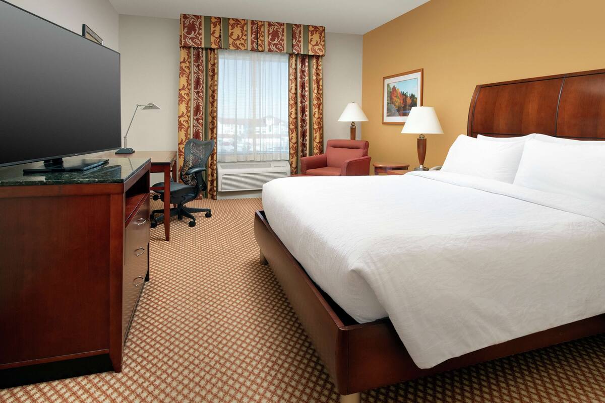Una habitación de hotel con una cama grande, un escritorio con televisor de pantalla plana, una silla, una lámpara y una ventana con cortinas.