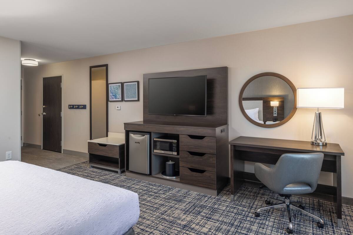 A hotel room with a bed, a desk with a chair, a television, a microwave, a small refrigerator, a trash can, and a mirror.