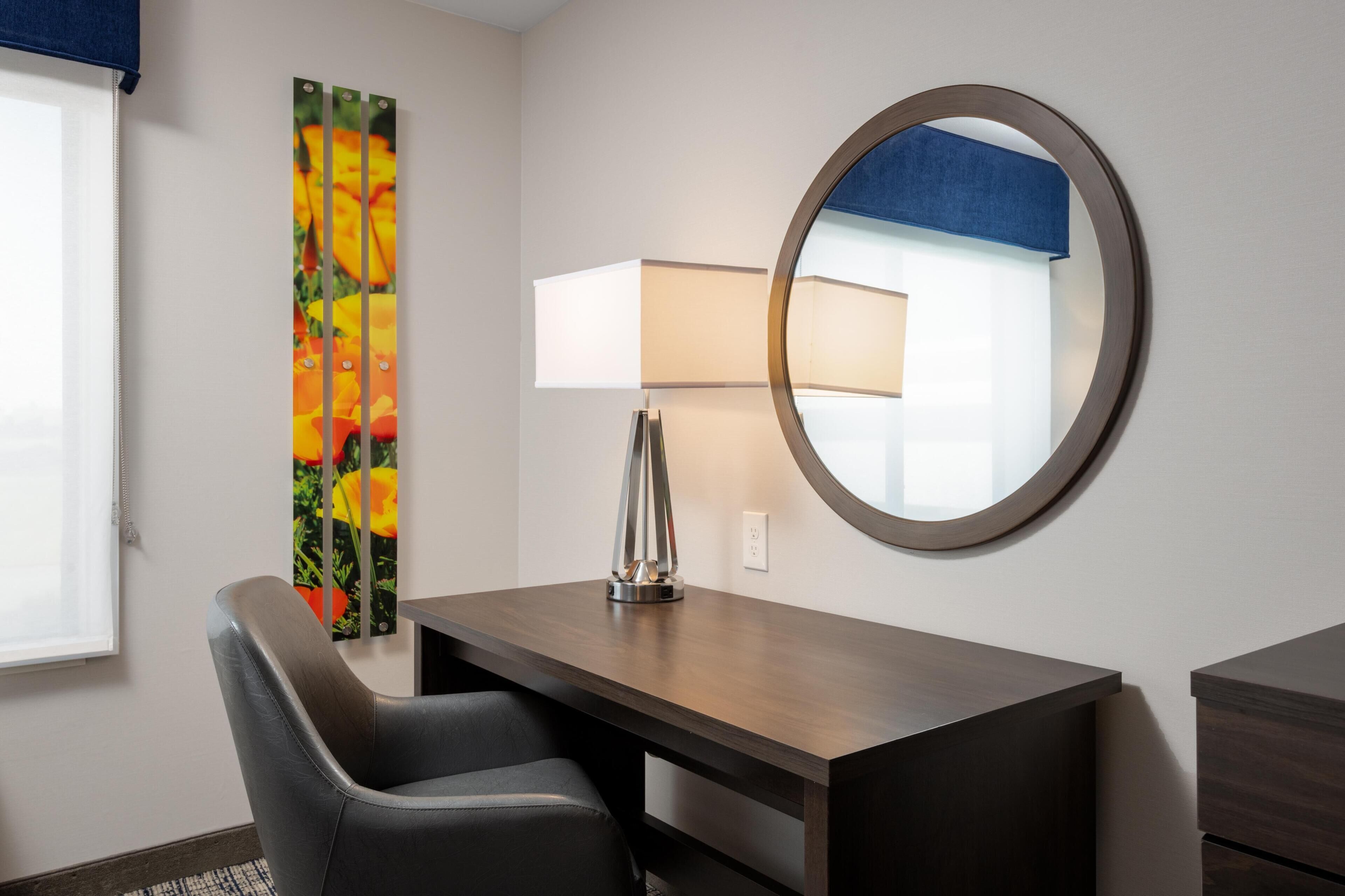 A hotel room with a desk, chair, lamp, mirror, and a colorful artwork on the wall.
