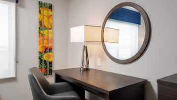 A hotel room with a desk, chair, lamp, mirror, and a colorful artwork on the wall.