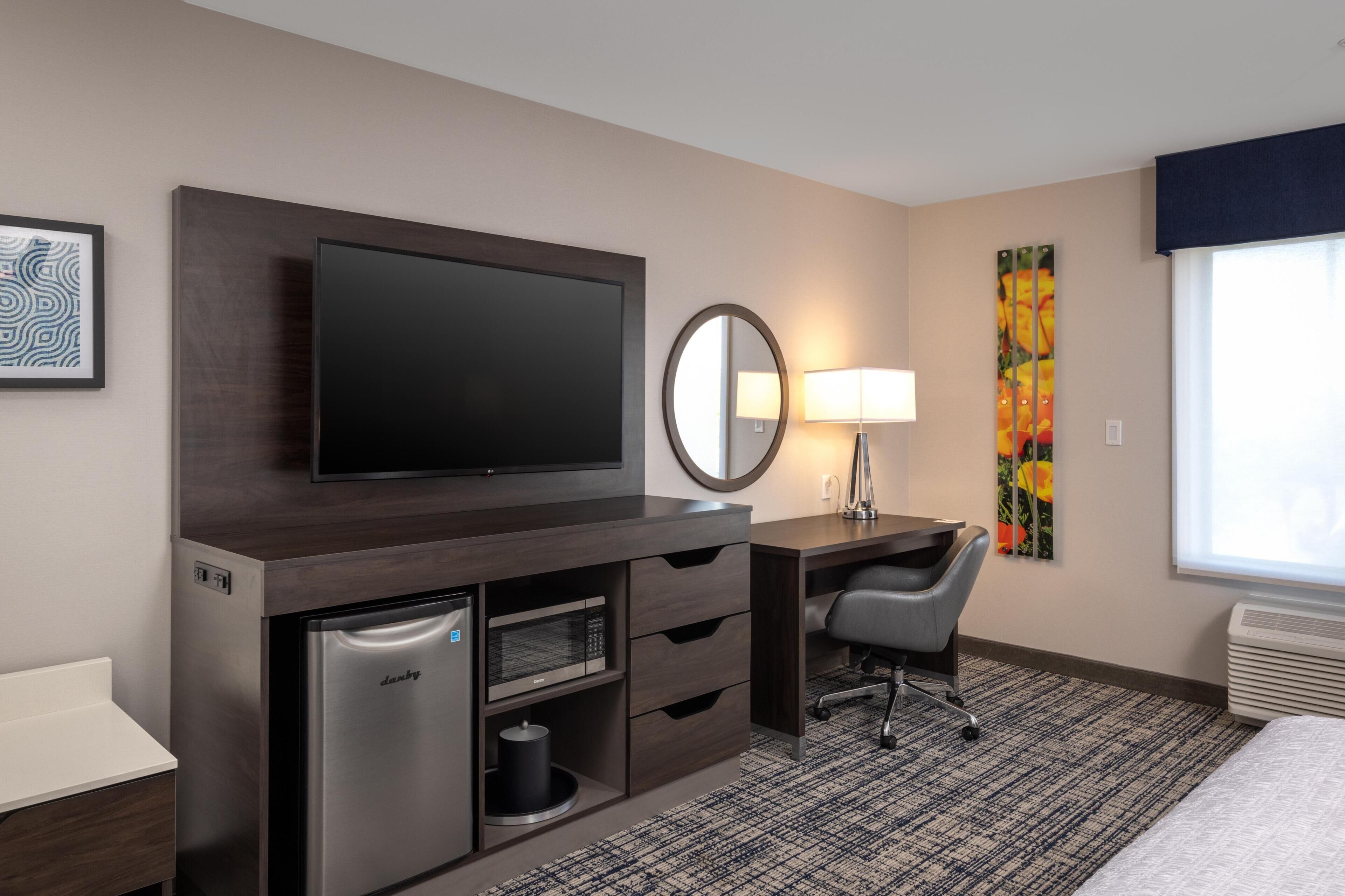 A hotel room with a flat-screen TV, a desk with a chair, a compact refrigerator, a microwave, a bedside table, a lamp, a large window, and a patterned rug.