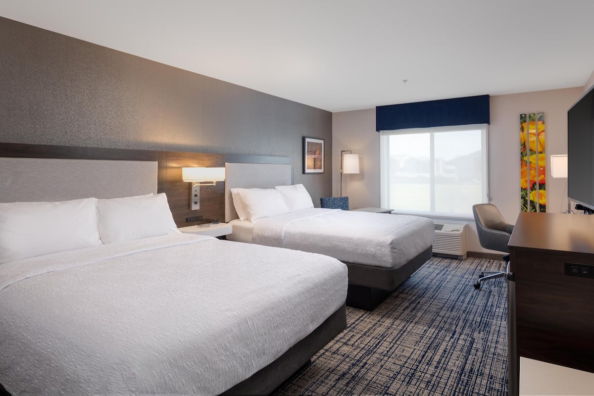 A hotel room with two beds, a desk, a chair, a large window, and a wall-mounted artwork.