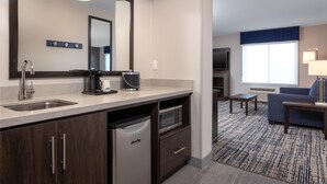 A modern hotel room with a kitchen area, a large mirror, a microwave, a dishwasher, a seating area with a sofa and a table, and a window with a view. - Hampton Inn & Suites Manteca (Manteca)