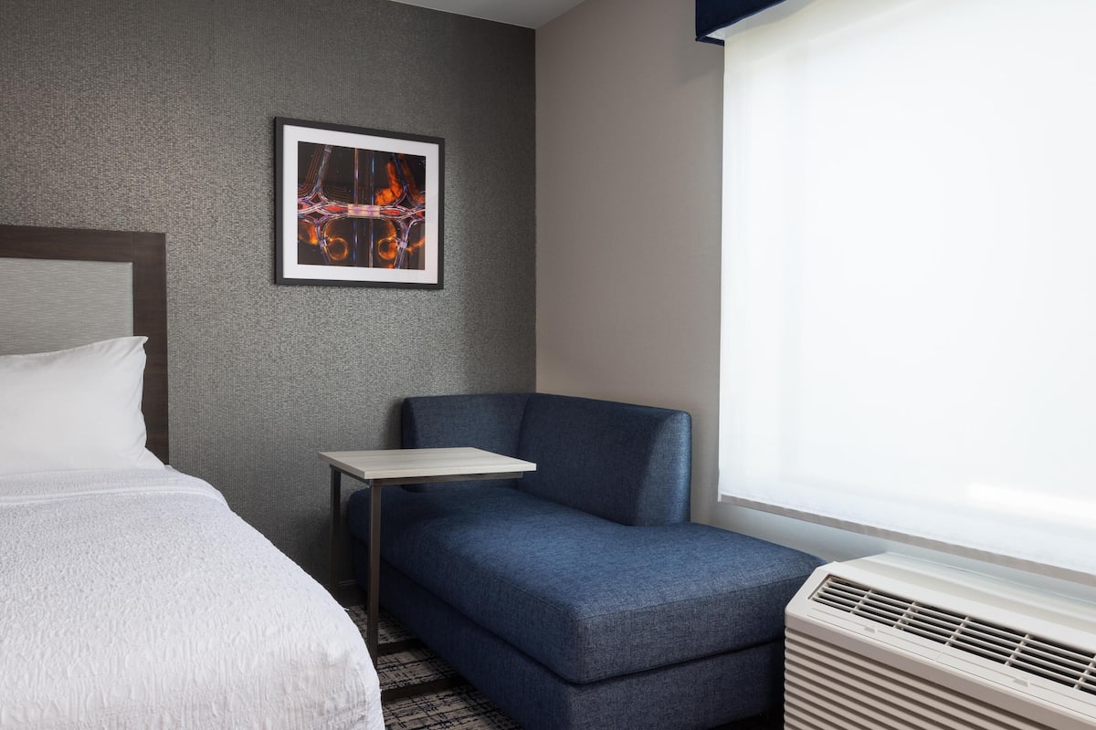 A hotel room with a bed, a small table, a blue armchair, a heater, and a framed abstract artwork on the wall.