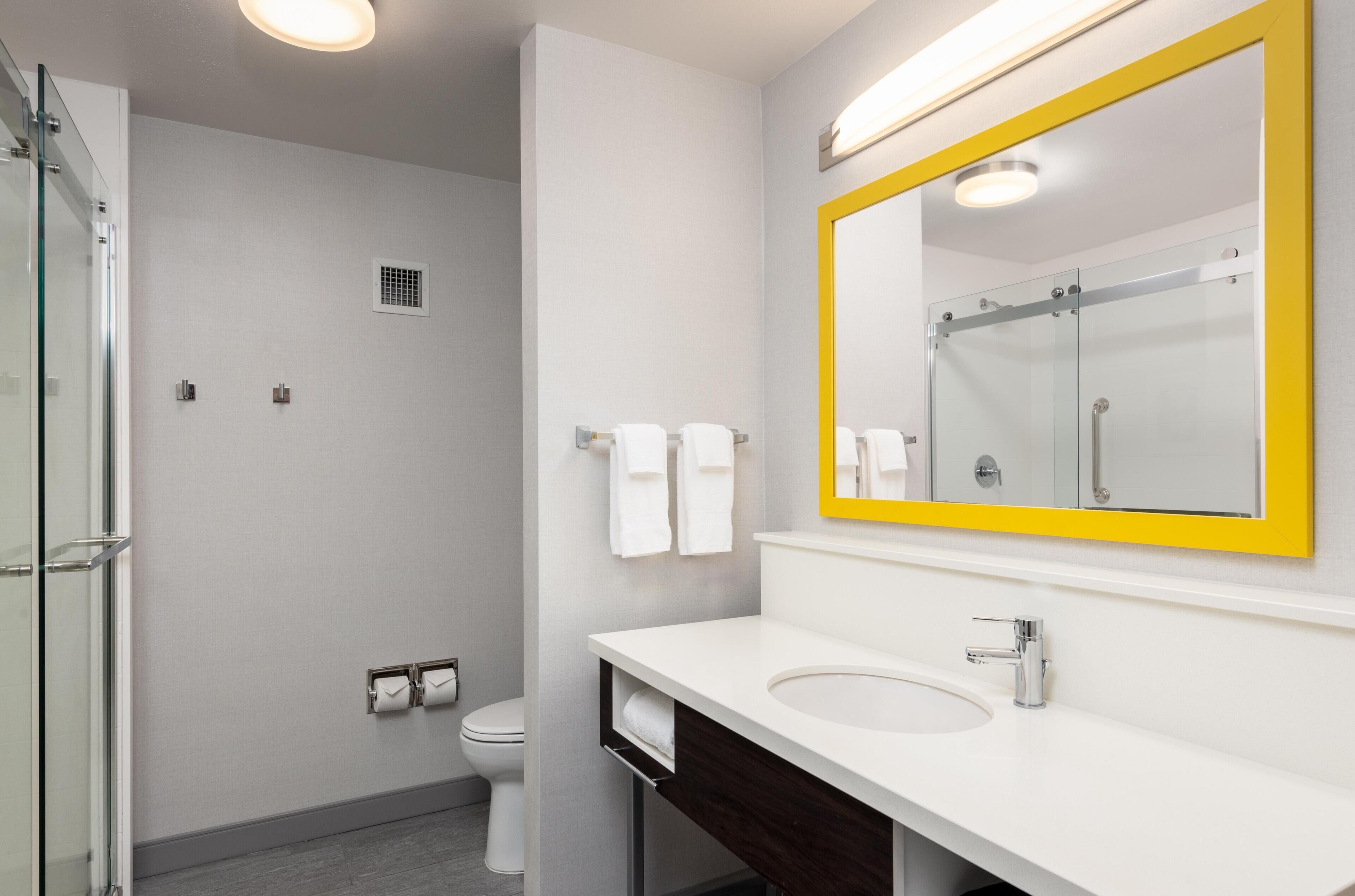 A modern bathroom with a glass shower enclosure, a white sink, and a large mirror with a yellow frame.