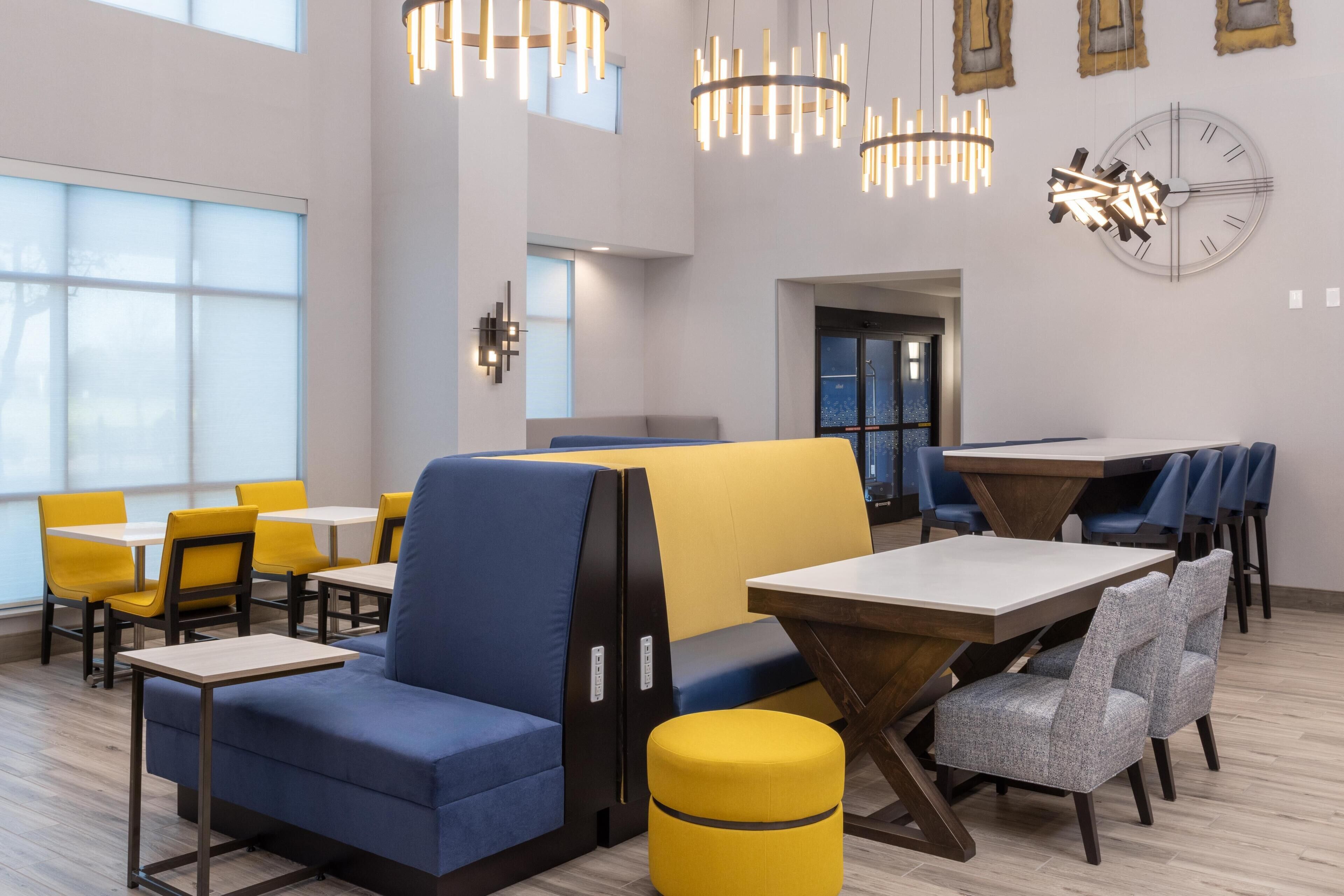 A modern dining area with high tables, blue and yellow seating, and pendant lighting.