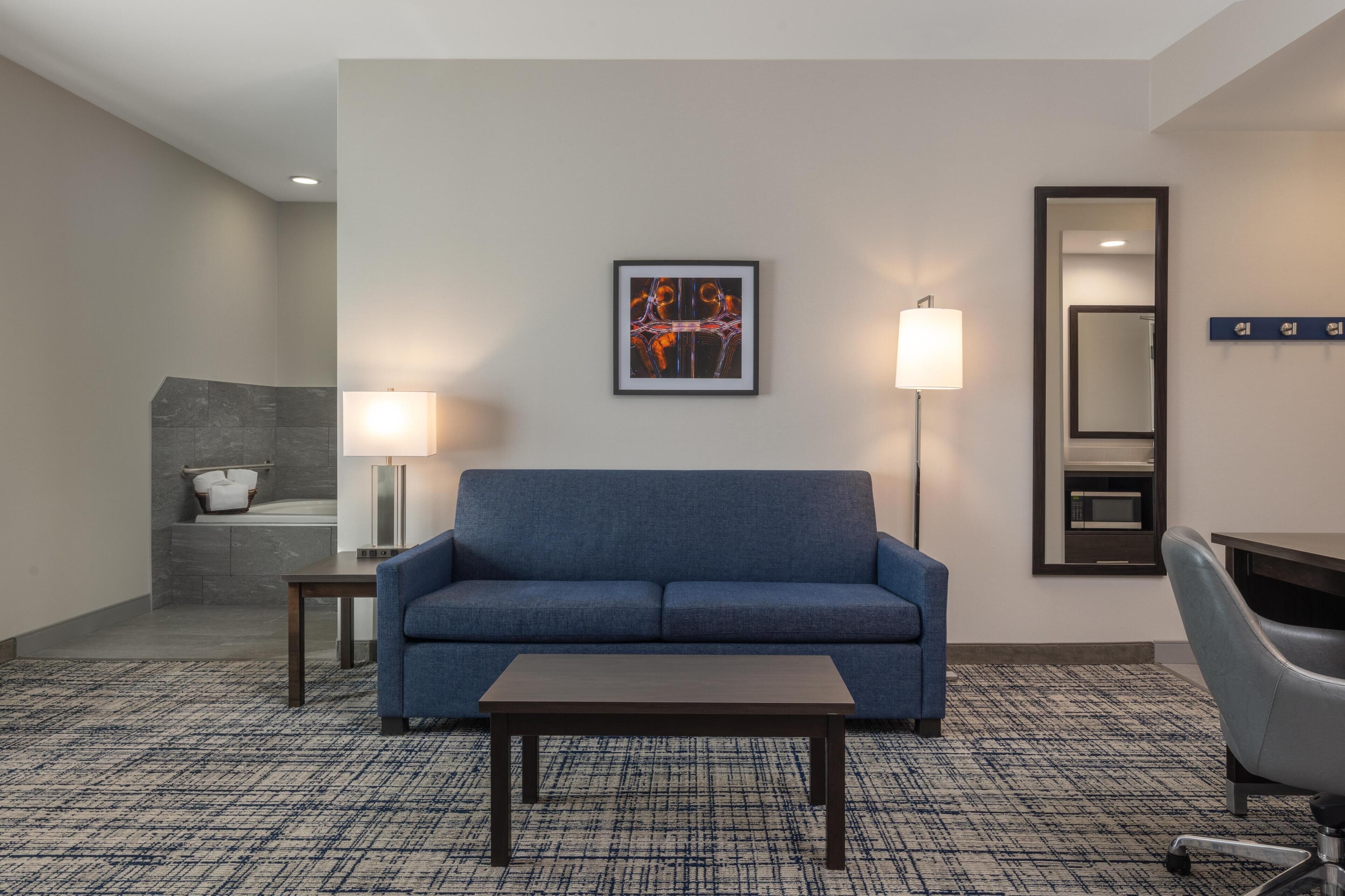 A hotel room with a blue sofa, a small table, a lamp, a mirror, and a framed artwork on the wall.