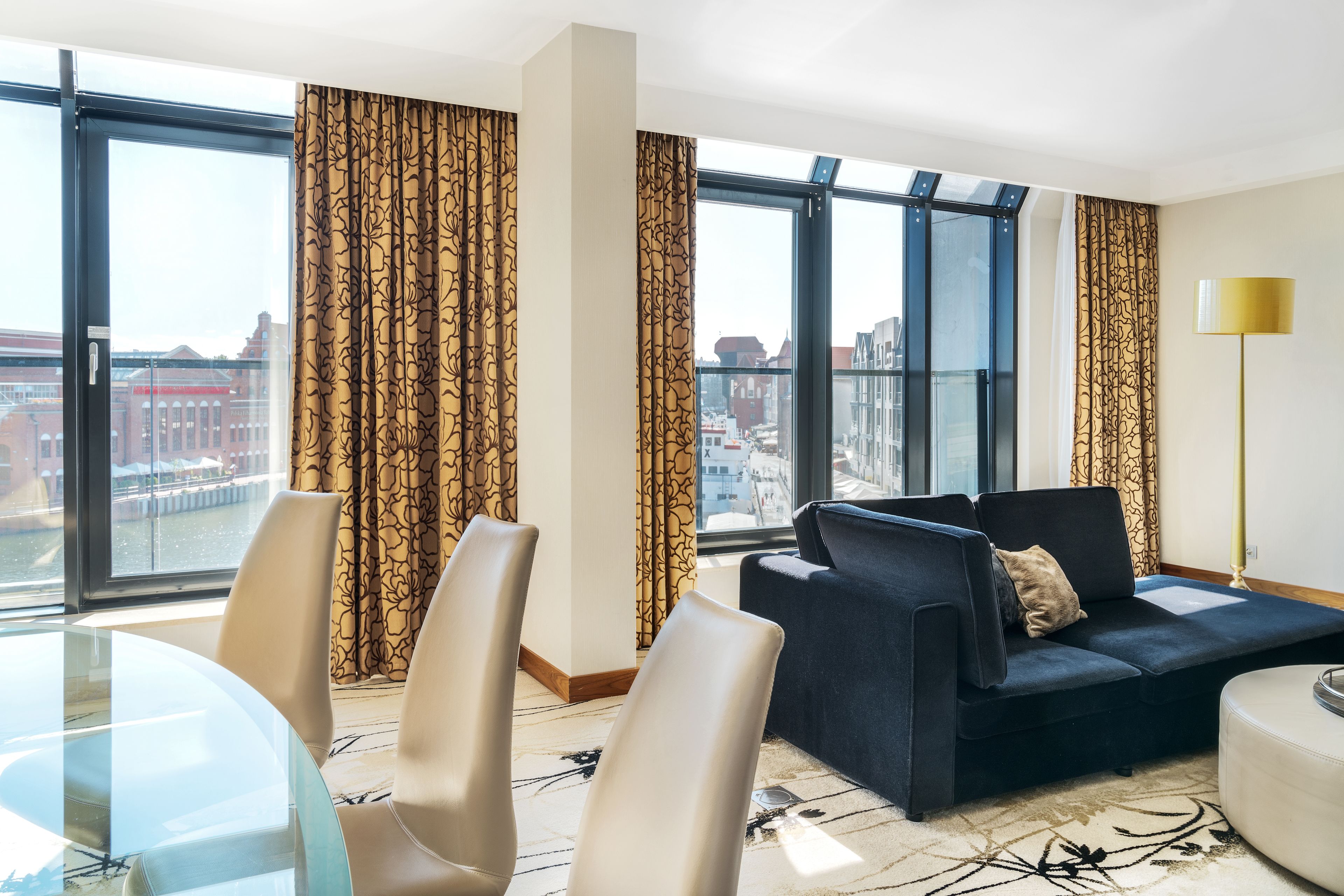 A modern hotel room with a glass dining table, two sofas, and a large window offering a city view.