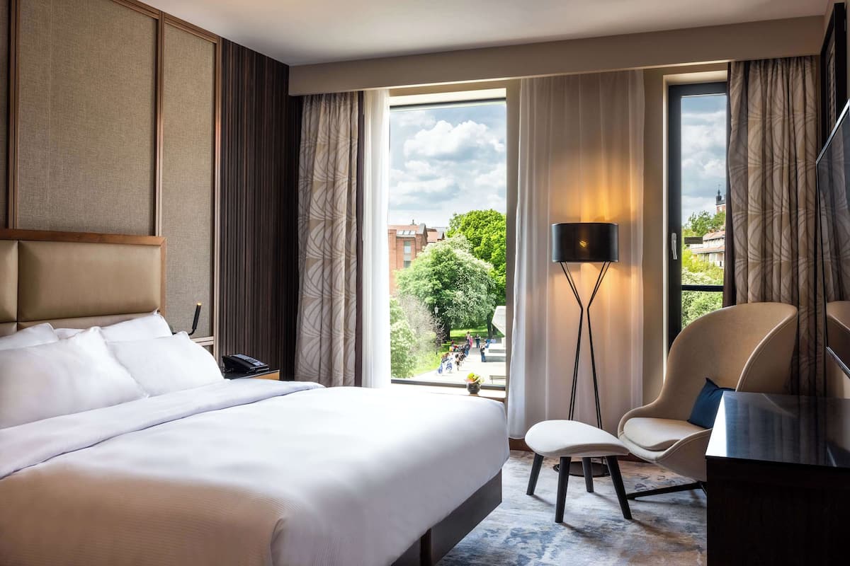 Quarto de hotel com uma cama grande, uma escrivaninha com cadeira, um abajur e vista para a cidade através de amplas janelas.