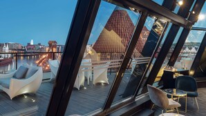 A modern rooftop bar with glass walls, offering a view of a cityscape and a distinctive red-roofed building.