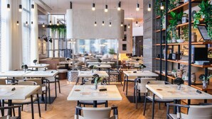 A modern restaurant interior with multiple tables and chairs, hanging lights, and a view of the city through large windows.