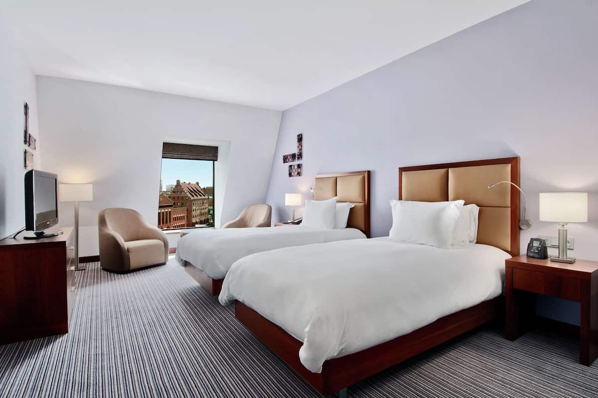 Quarto de hotel com duas camas, televisão, uma cadeira e janela com vista para a cidade.