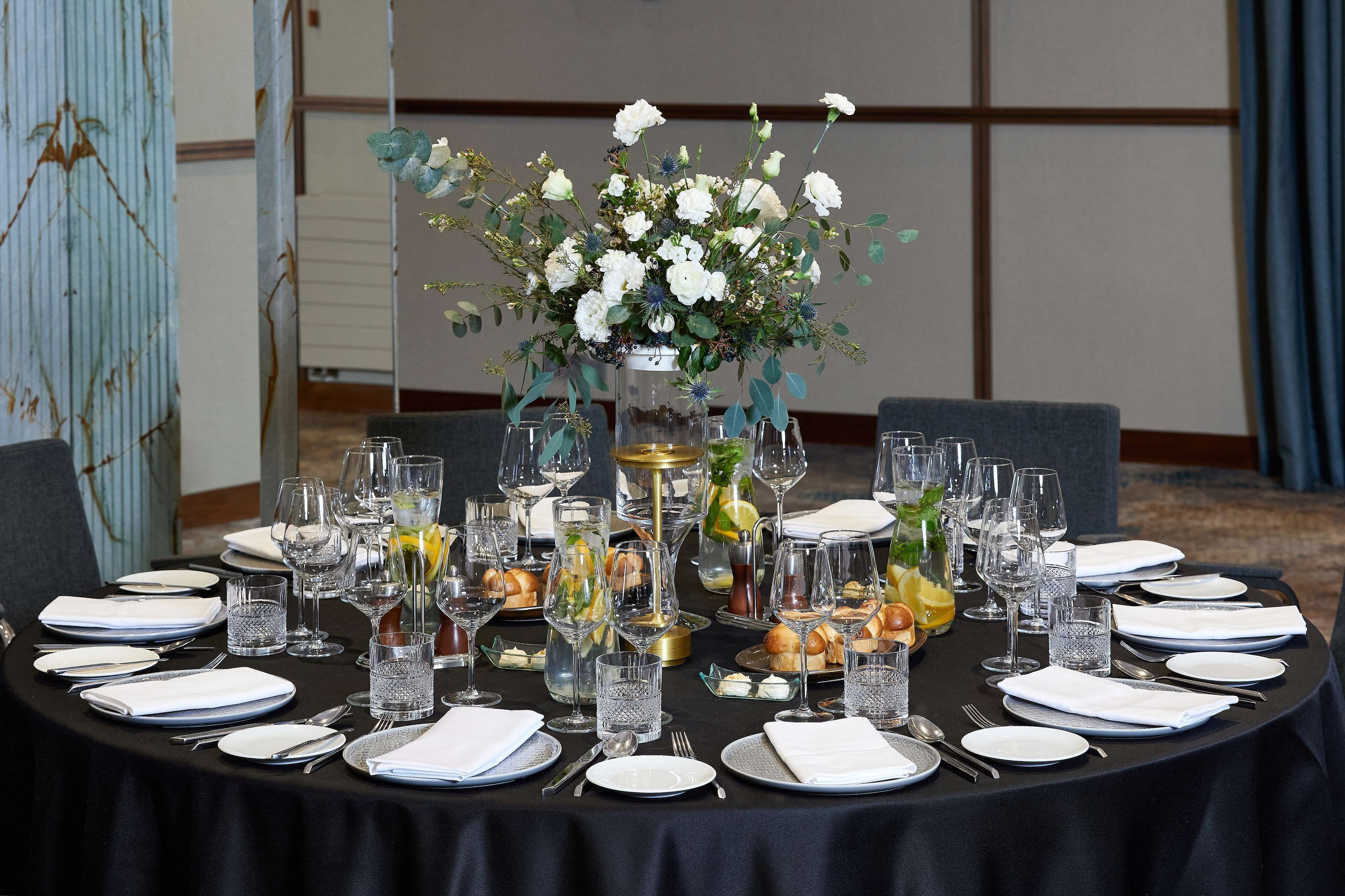 A round table set for a formal dinner with white plates, silverware, and clear glassware. A centerpiece with white flowers and greenery is placed in the middle.