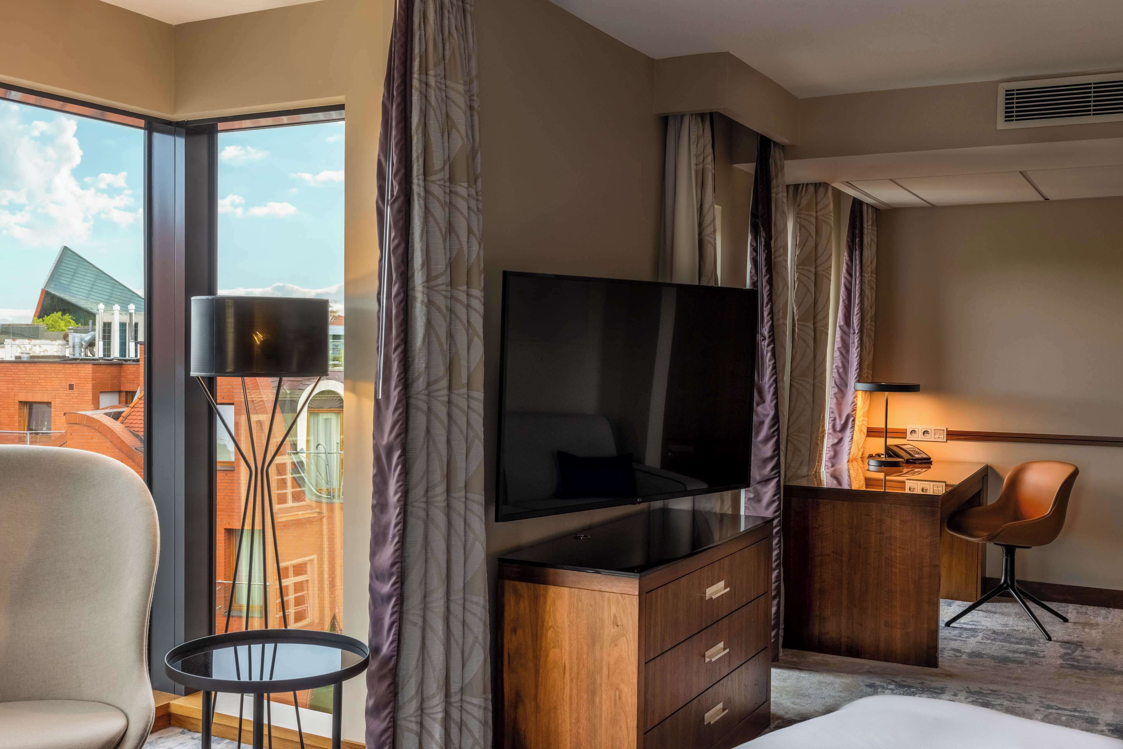Habitación de hotel con televisor grande, escritorio, silla y vistas a edificios desde la ventana.