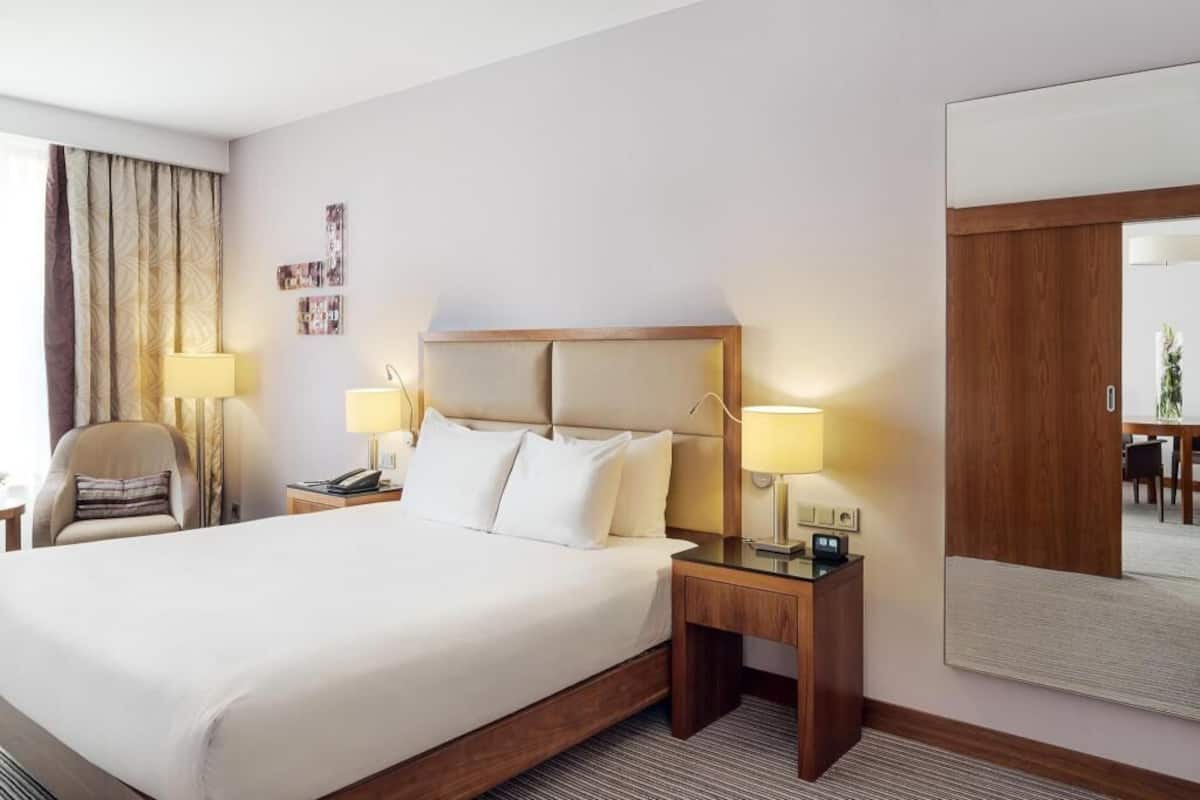 Quarto de hotel com uma cama grande, duas mesas de cabeceira com luminárias, uma cadeira, uma escrivaninha e um espelho grande.
