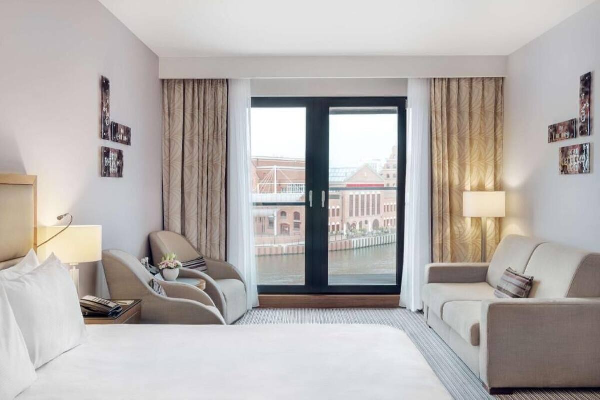 Quarto de hotel com uma cama, duas poltronas, um sofá, uma janela grande com vista para edifícios e uma porta.