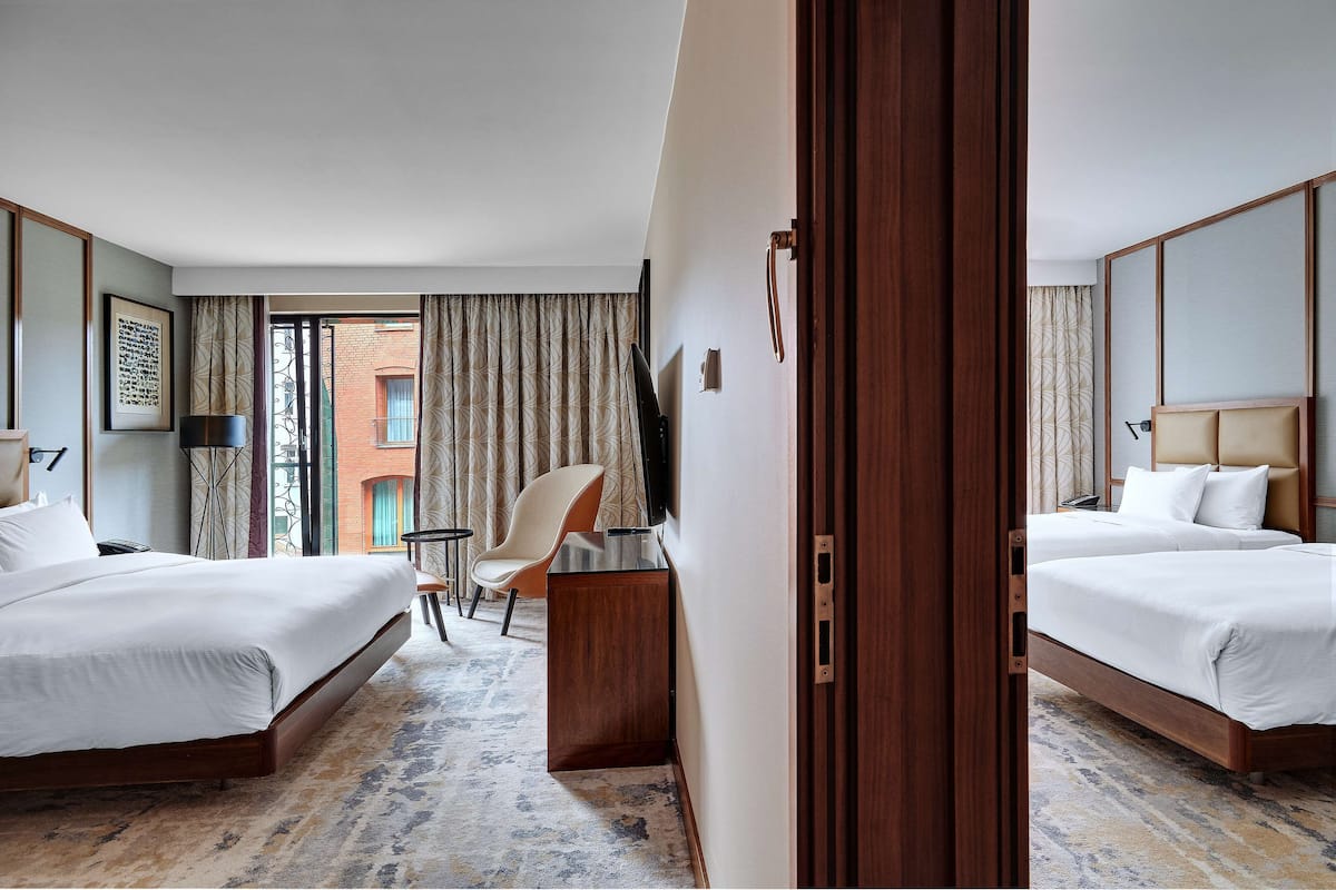Quarto de hotel com duas camas, uma escrivaninha, uma cadeira e uma janela grande com cortinas.