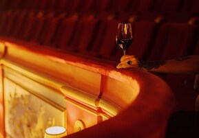 Primer plano de una copa de vino sobre un repisa de madera, con un fondo borroso de asientos rojos y una pared con un diseño circular.