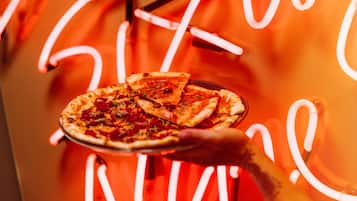 Een hand houdt een pizza met toppings vast, voor een neonbord.