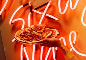 Una mano sosteniendo una pizza con ingredientes, frente a un letrero de neón.