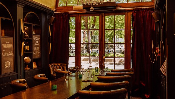 A dining area with a long table, multiple chairs, a chandelier, and large windows with curtains.