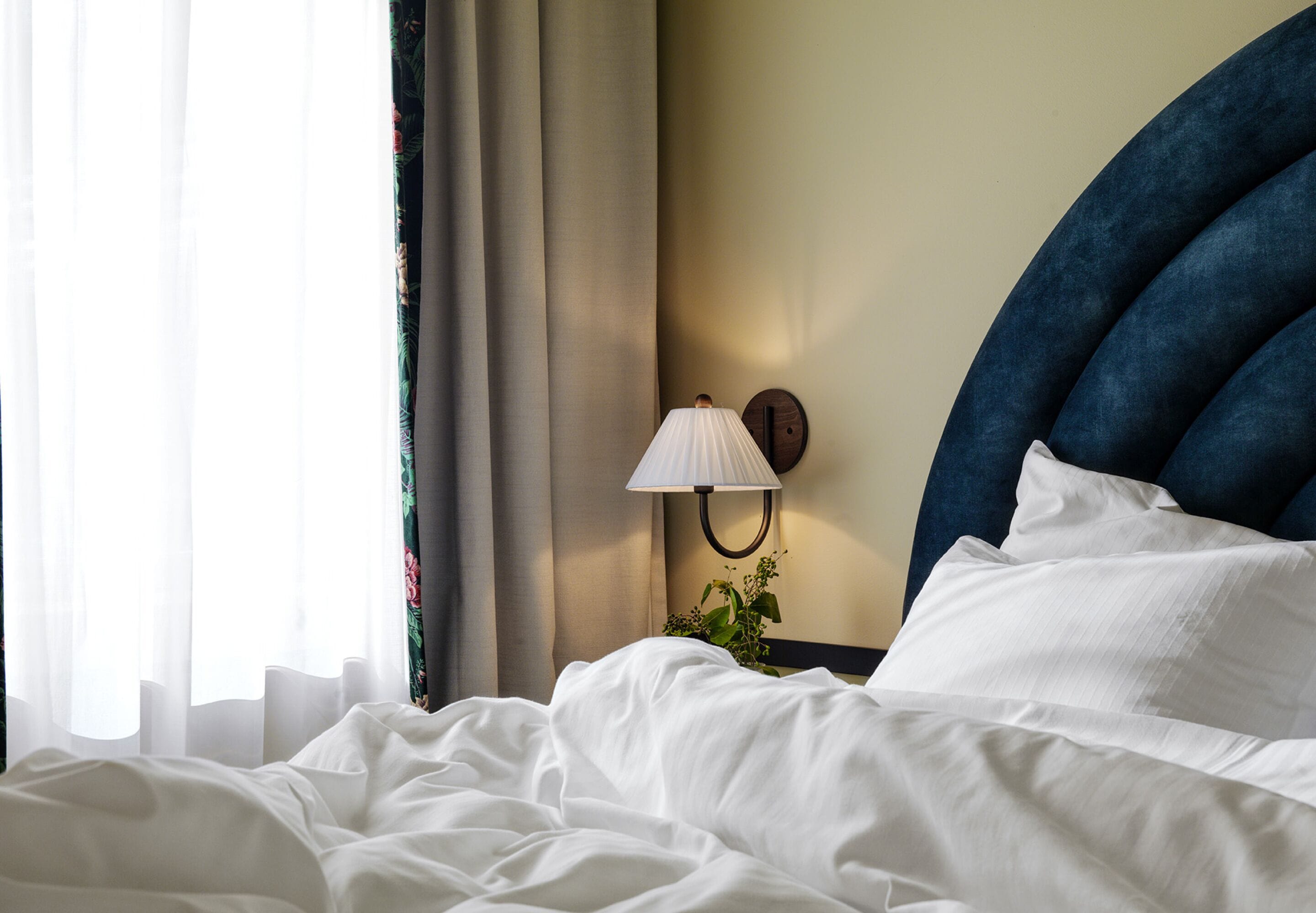 Ein ordentlich bezogenes Bett mit weißen Leinen, einer an der Wand befestigten Lampe und einem dunkelblauen Kopfteil.