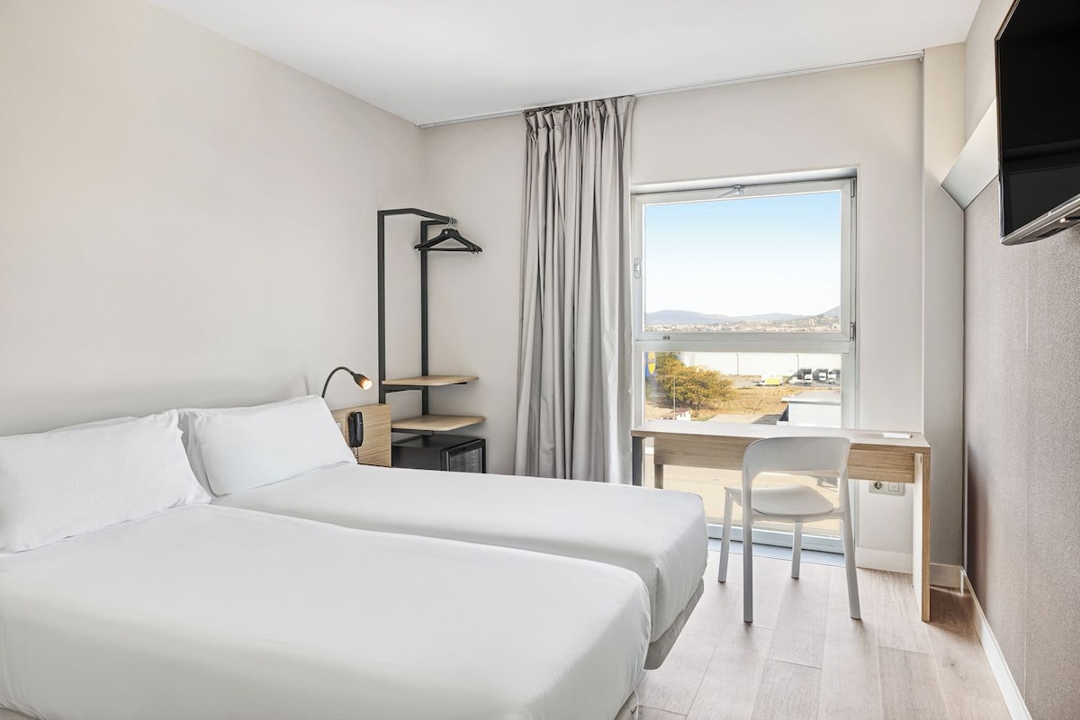 A hotel room with two beds, a desk, a chair, a lamp, and a view of the outdoors.