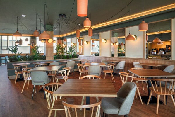 A modern restaurant interior with wooden tables and chairs, pendant lighting, and large windows allowing natural light. - B&B Hotel Granada (Pulianas)