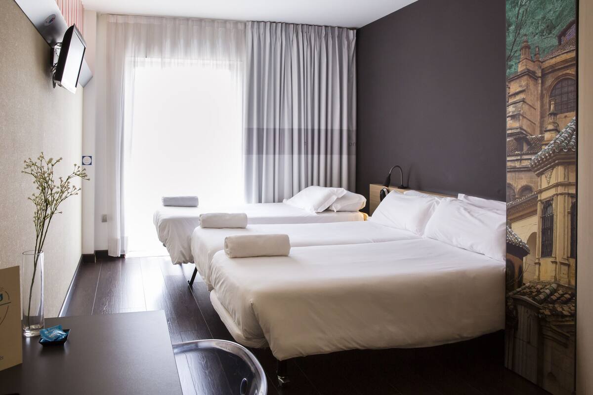 A hotel room with two beds, a desk, a chair, a vase with flowers, and a painting on the wall.