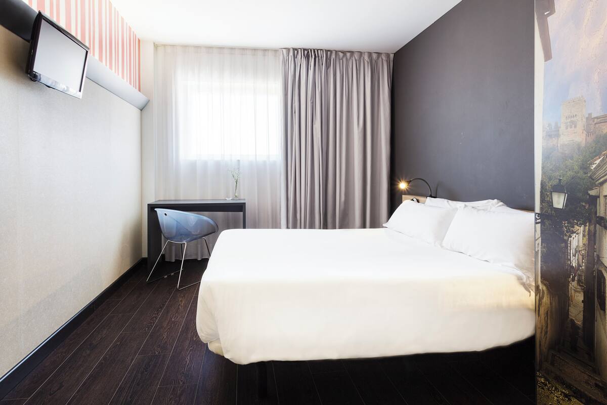 A hotel room with a bed, a desk, a chair, and a large window with curtains.