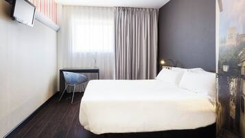 Quarto de hotel com uma cama, uma escrivaninha, uma cadeira e uma janela grande com cortinas.