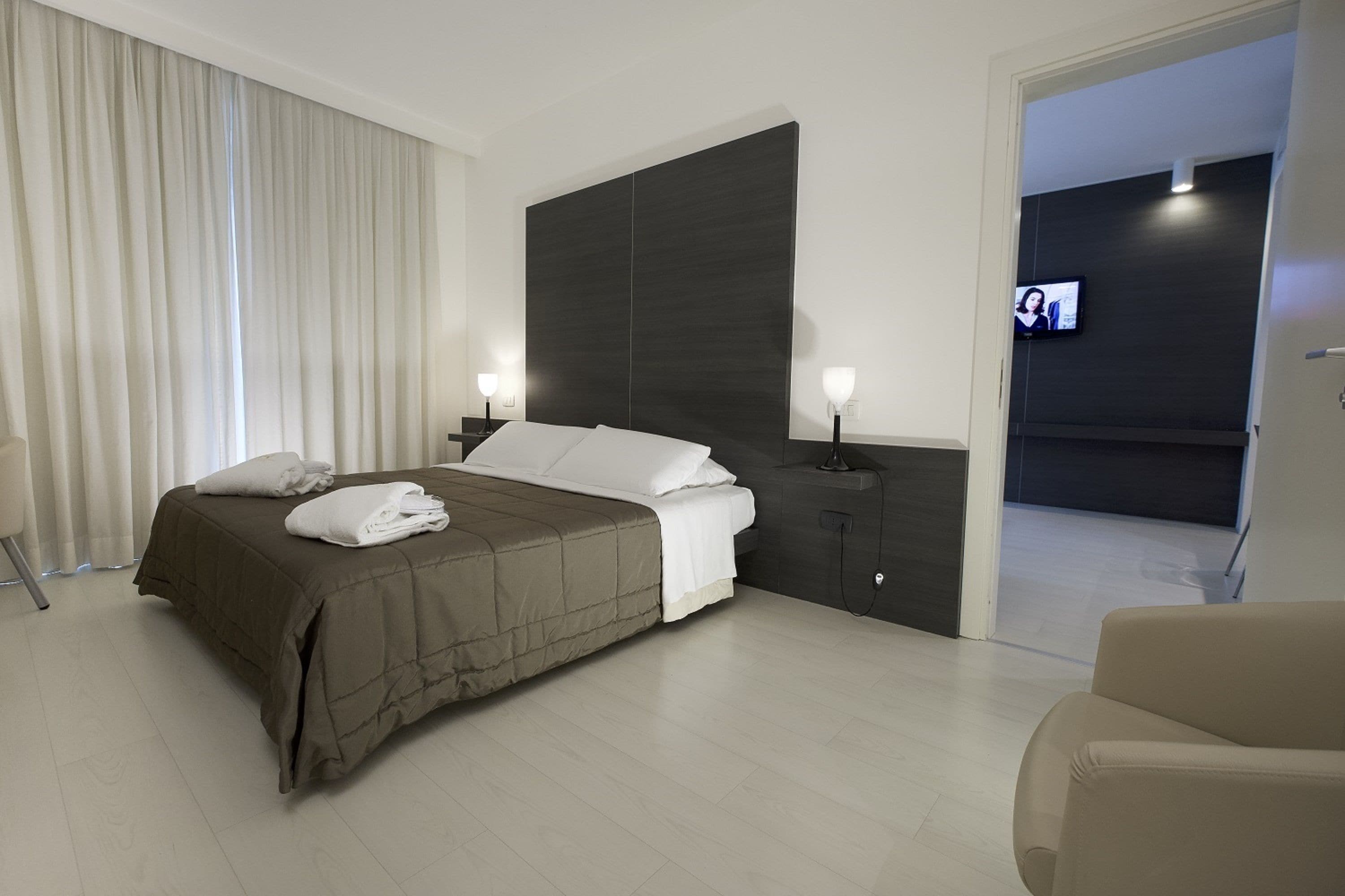 Ein modernes Hotelzimmer mit einem großen Bett, einem dunklen Kopfteil, zwei Nachttischlampen und einem an der Wand befestigten Fernseher.