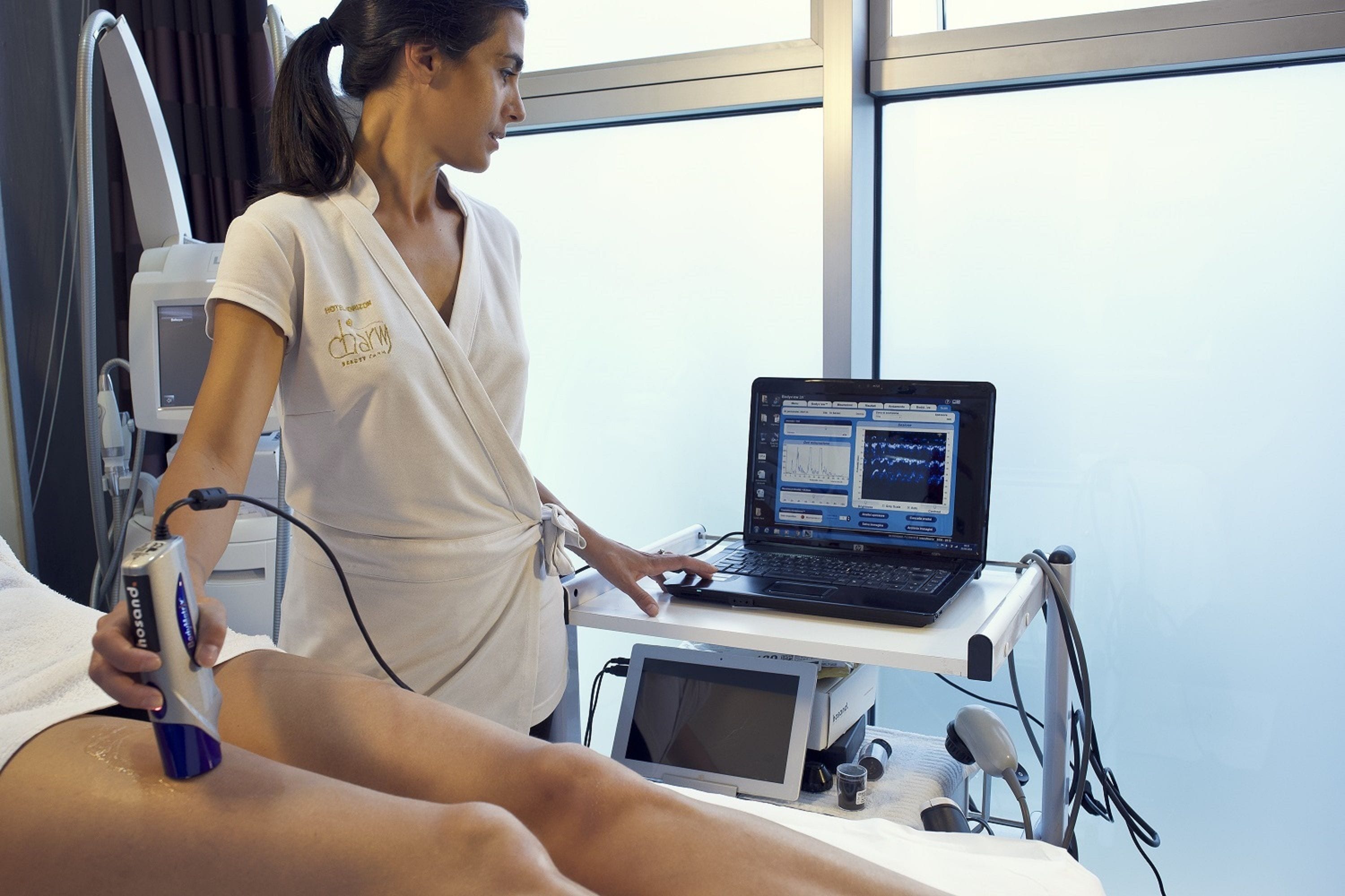 A medical professional using a device on a patient in a clinical setting with a laptop and medical equipment.