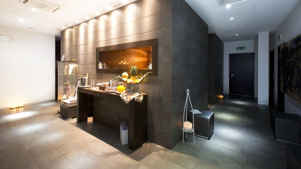 Un área de recepción moderna con un mostrador oscuro, una vitrina de cristal y un pequeño espacio de descanso con una silla y un cubo blanco.
