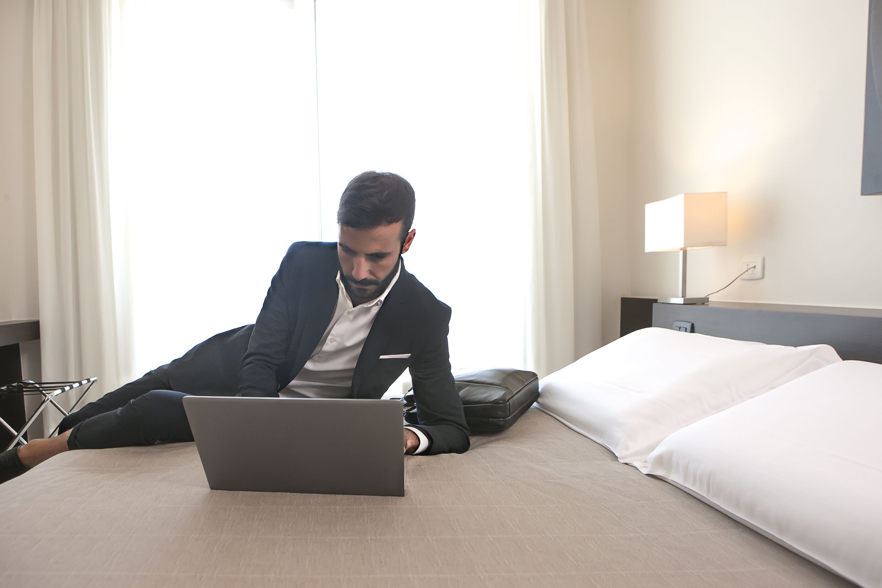 Un hombre con traje trabaja en una computadora portátil en una habitación de hotel, con una cama, almohadas y una mesita de noche con una lámpara.