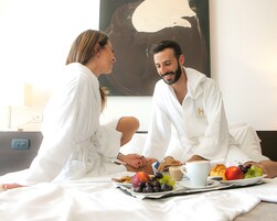 A couple sitting on a bed with breakfast, including fruits, coffee, and pastries.