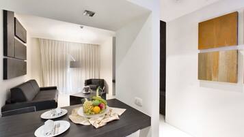 Una habitación moderna y bien iluminada con una zona de comedor que incluye una mesa de madera oscura, platos blancos y una bandeja de frutas. Hay un sofá negro y un gran ventanal con cortinas.