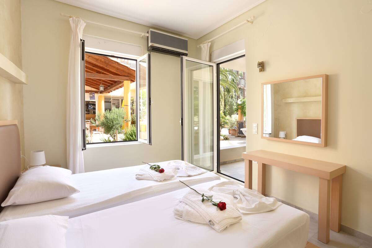 A hotel room with two beds, a large window, a sliding glass door, a wooden bench, and a mirror.