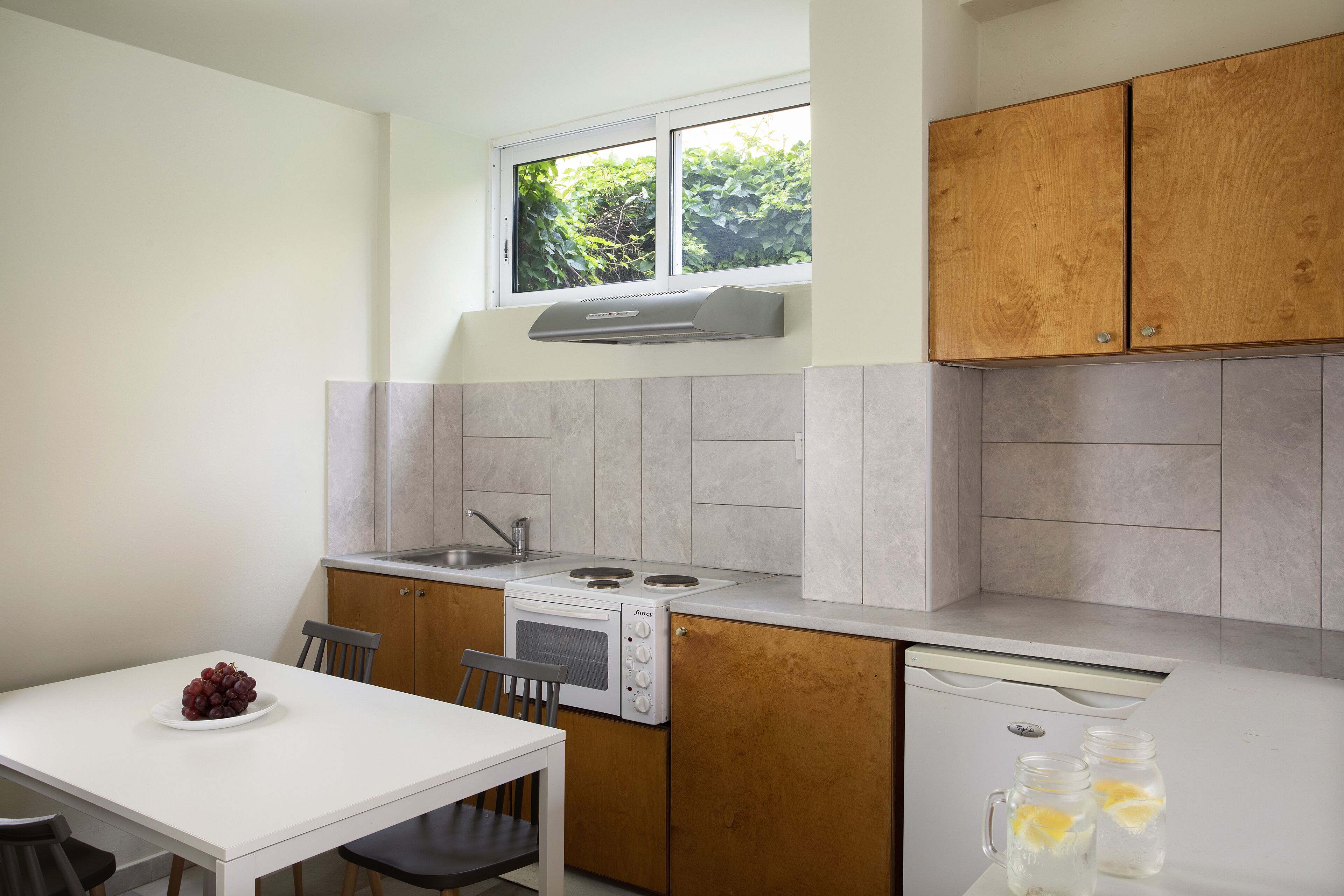 A compact kitchen with a built-in oven, a microwave, and a sink. There is a window above the sink, and a white table with chairs is positioned in front of the kitchen.