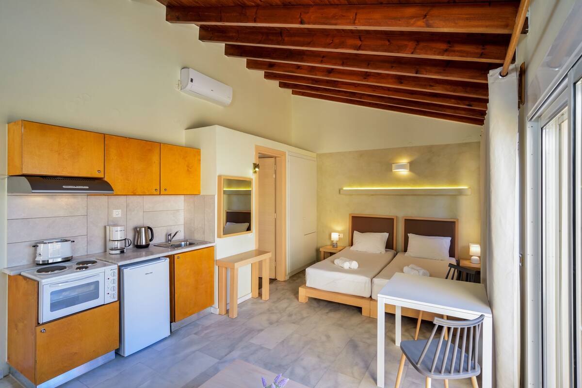 A compact hotel room with a kitchenette, a bed with two pillows, a bedside table with a lamp, and a small seating area.