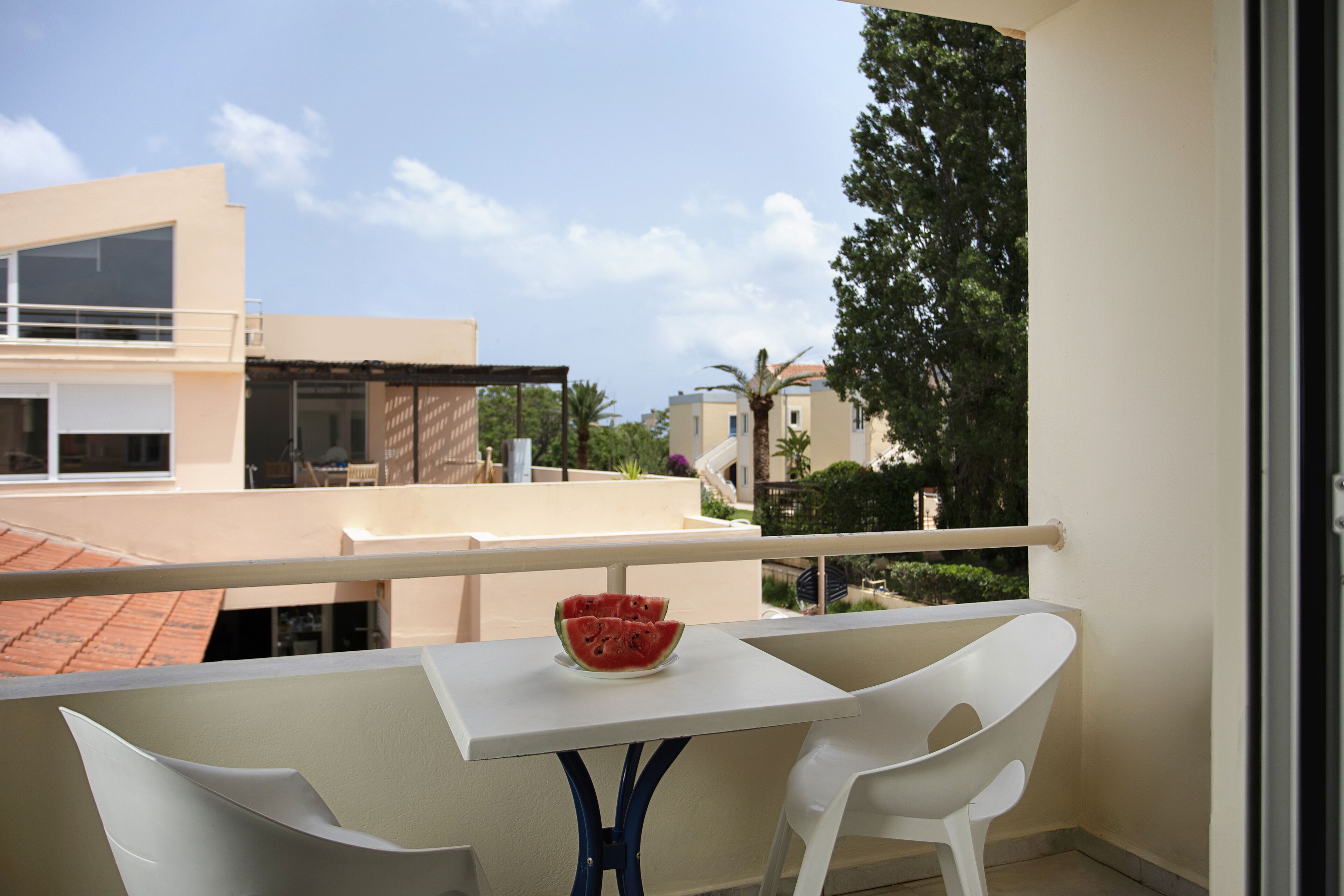A balcony with a white table and two white chairs, a bowl of watermelon, and a view of buildings and trees.