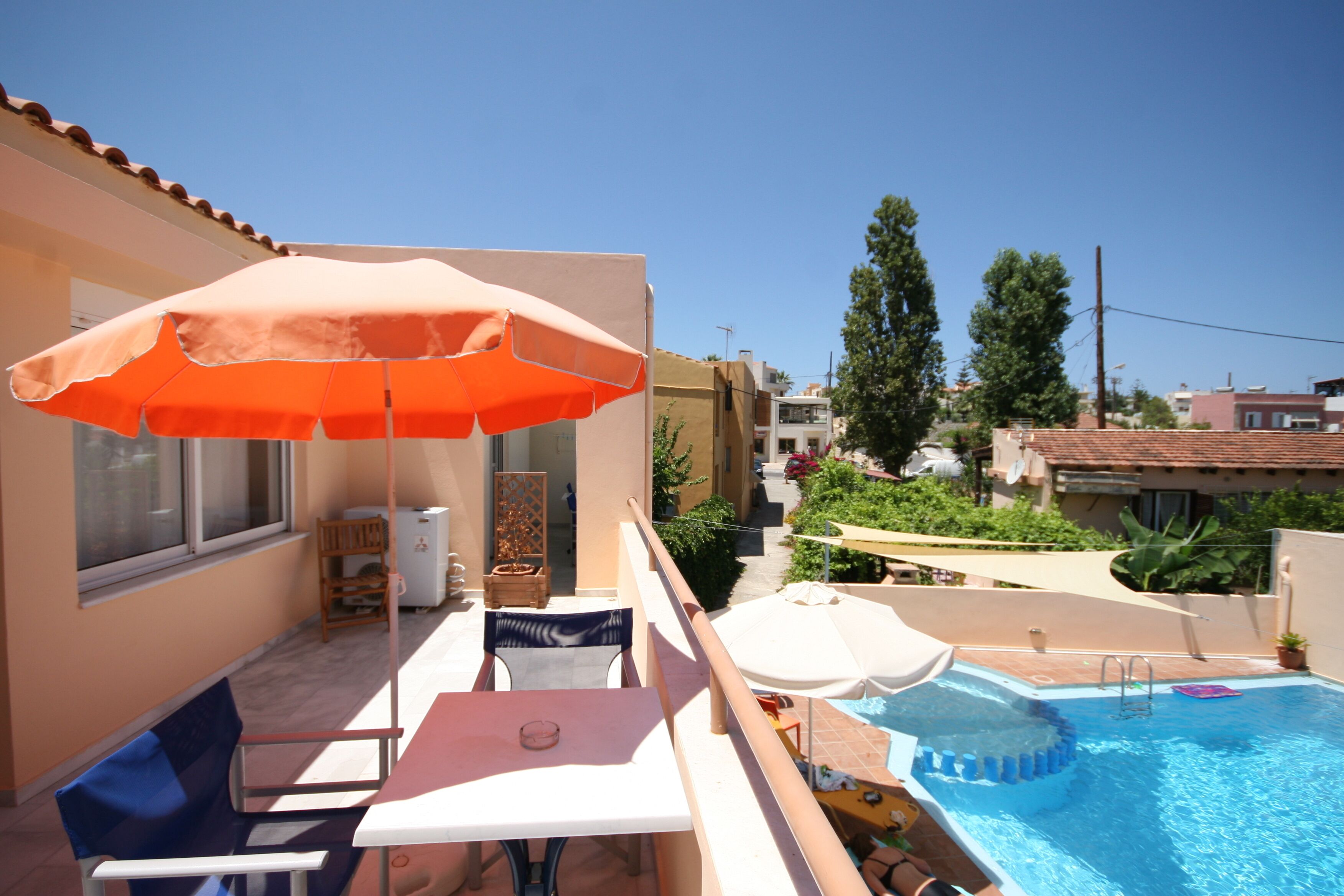 A balcony with a table, chairs, and an orange umbrella. There is a swimming pool with a ladder and a small deck area.