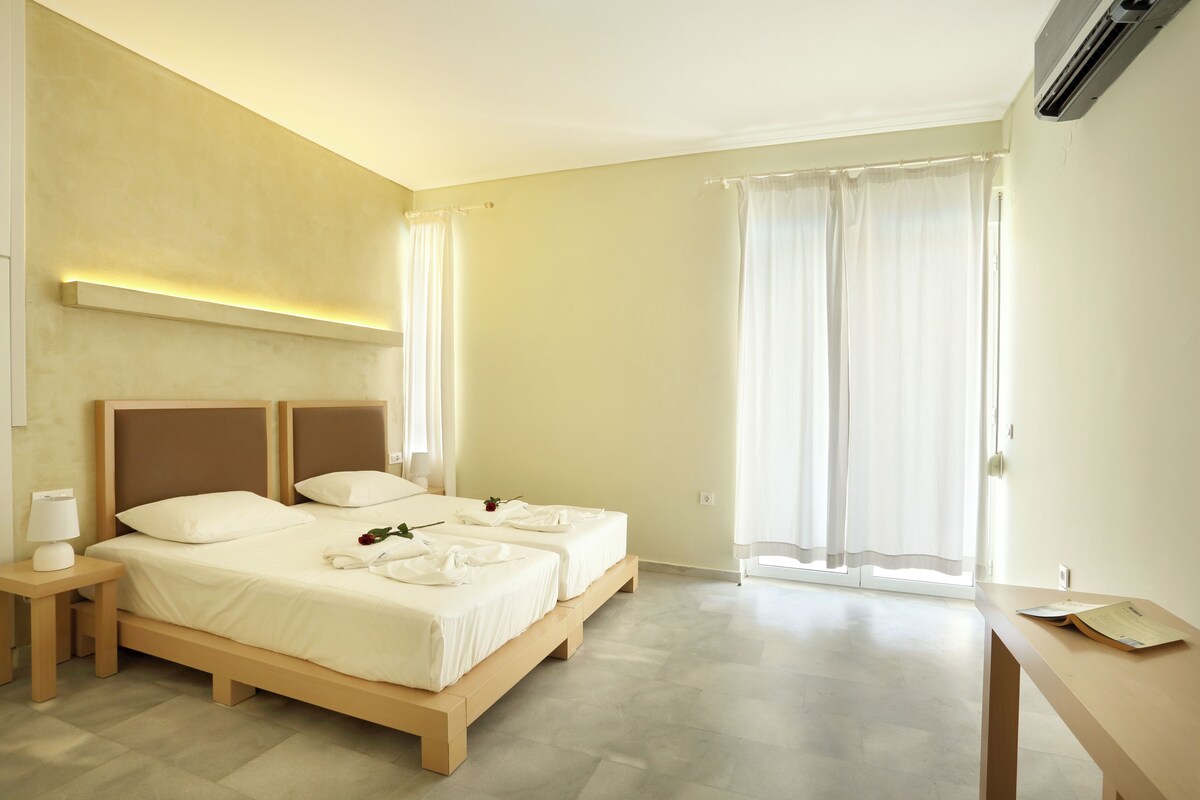 A hotel room with two beds, a bedside table with a lamp, a desk with a chair, and a large window with curtains.