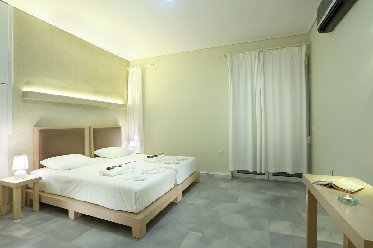 A hotel room with two beds, a bedside table with a lamp, a small table with a book and a candle, and a window with curtains.