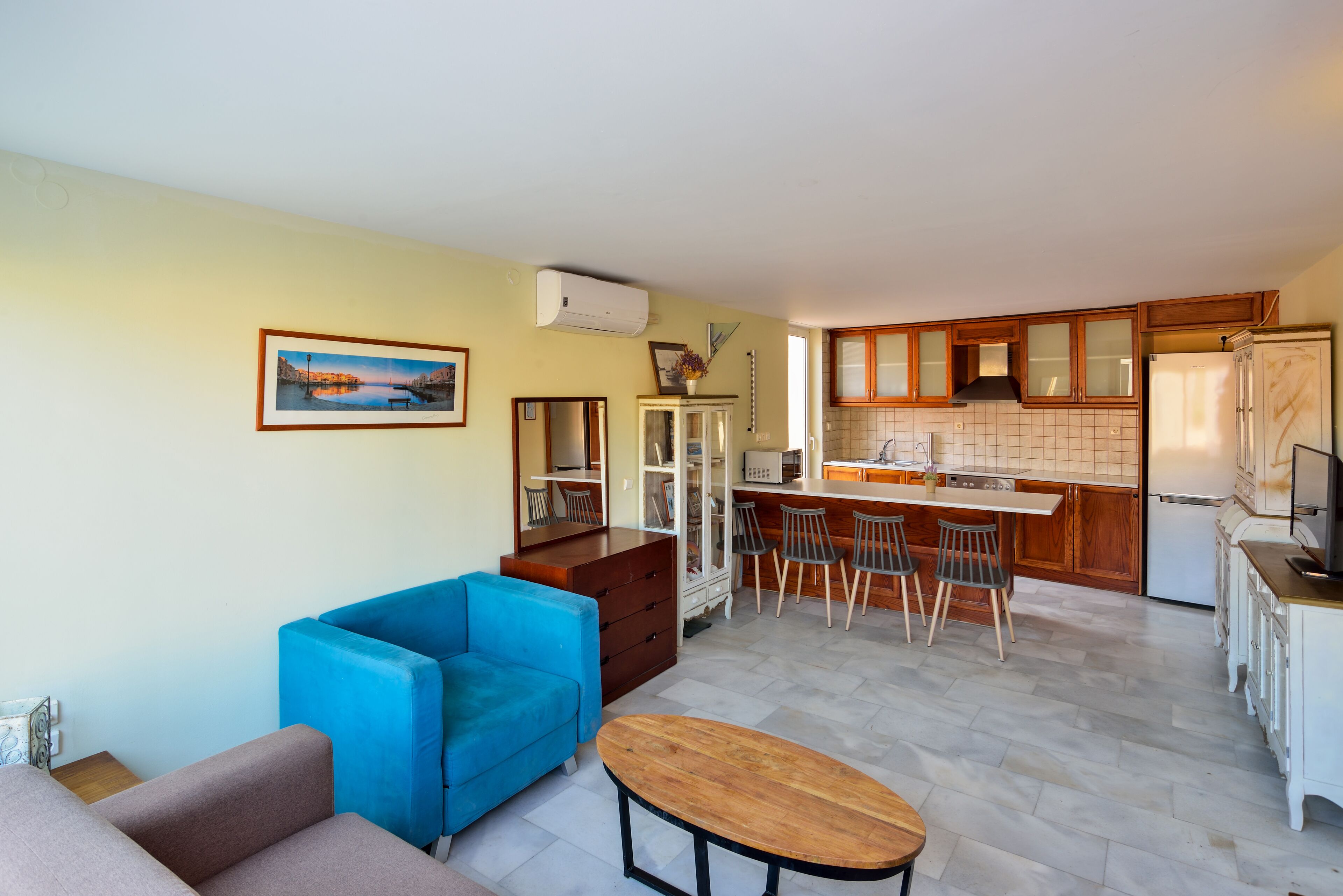 A compact living space with a blue sofa, a wooden coffee table, a kitchen area with wooden cabinets and a refrigerator, and a dining area with bar stools.