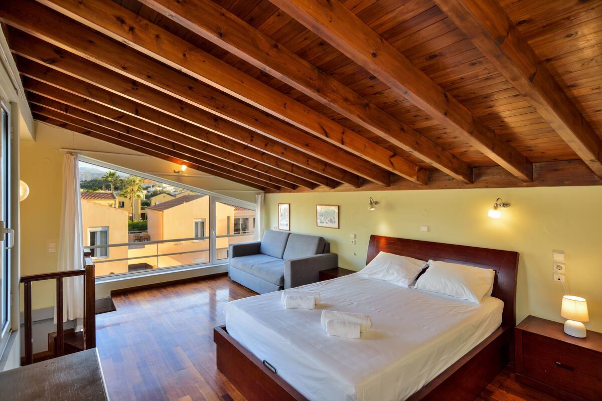 A bedroom with a wooden ceiling, a bed, a sofa, and a window with a view of buildings outside.