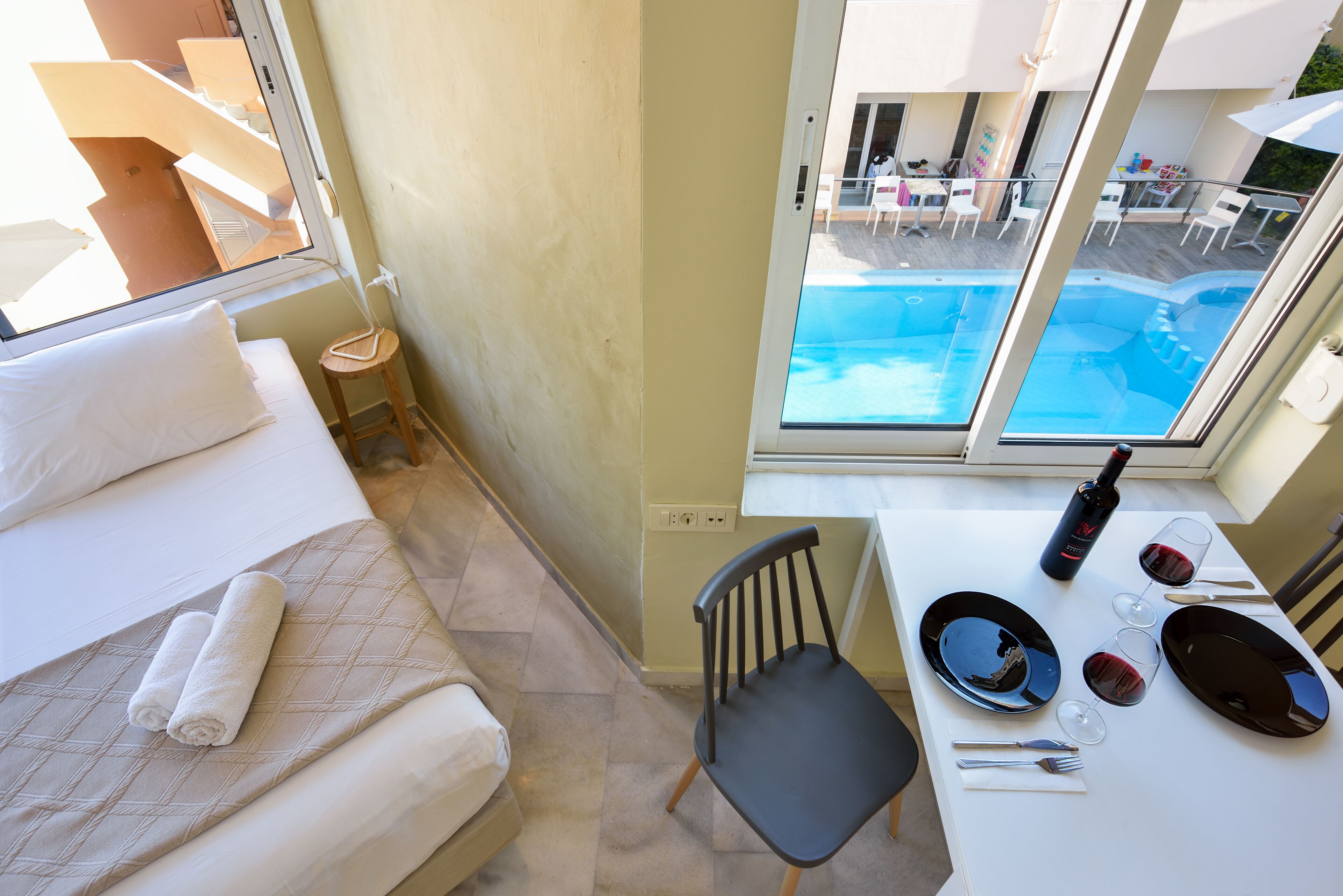 A hotel room with a bed, a small table with a bottle and glasses, and a view of a pool.