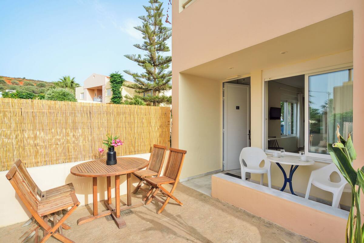 A patio with a round wooden table and chairs, a small white table with chairs, and a view of a building and trees in the background.